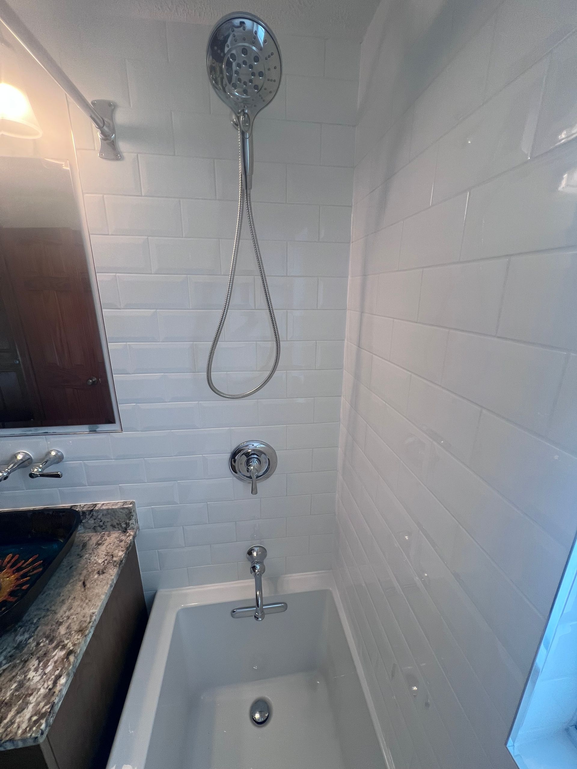 Small white bathroom shower stall with chrome showerhead, hose, and bathtub faucet beside a sink.