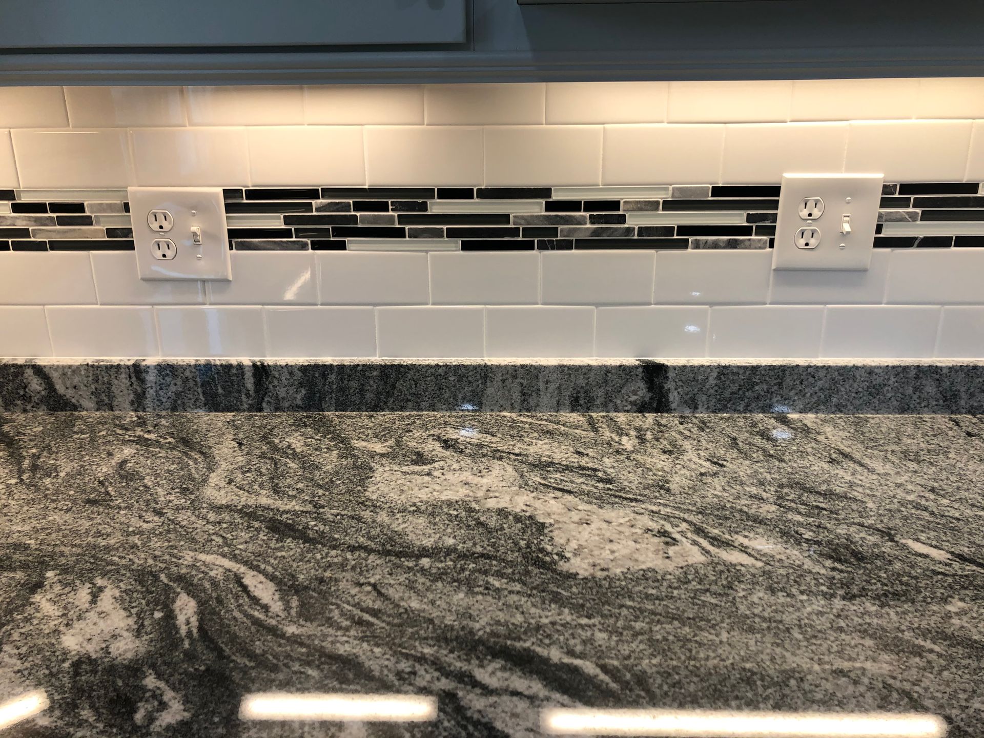 Kitchen backsplash with white tile, black accent strip, outlets, and granite countertop