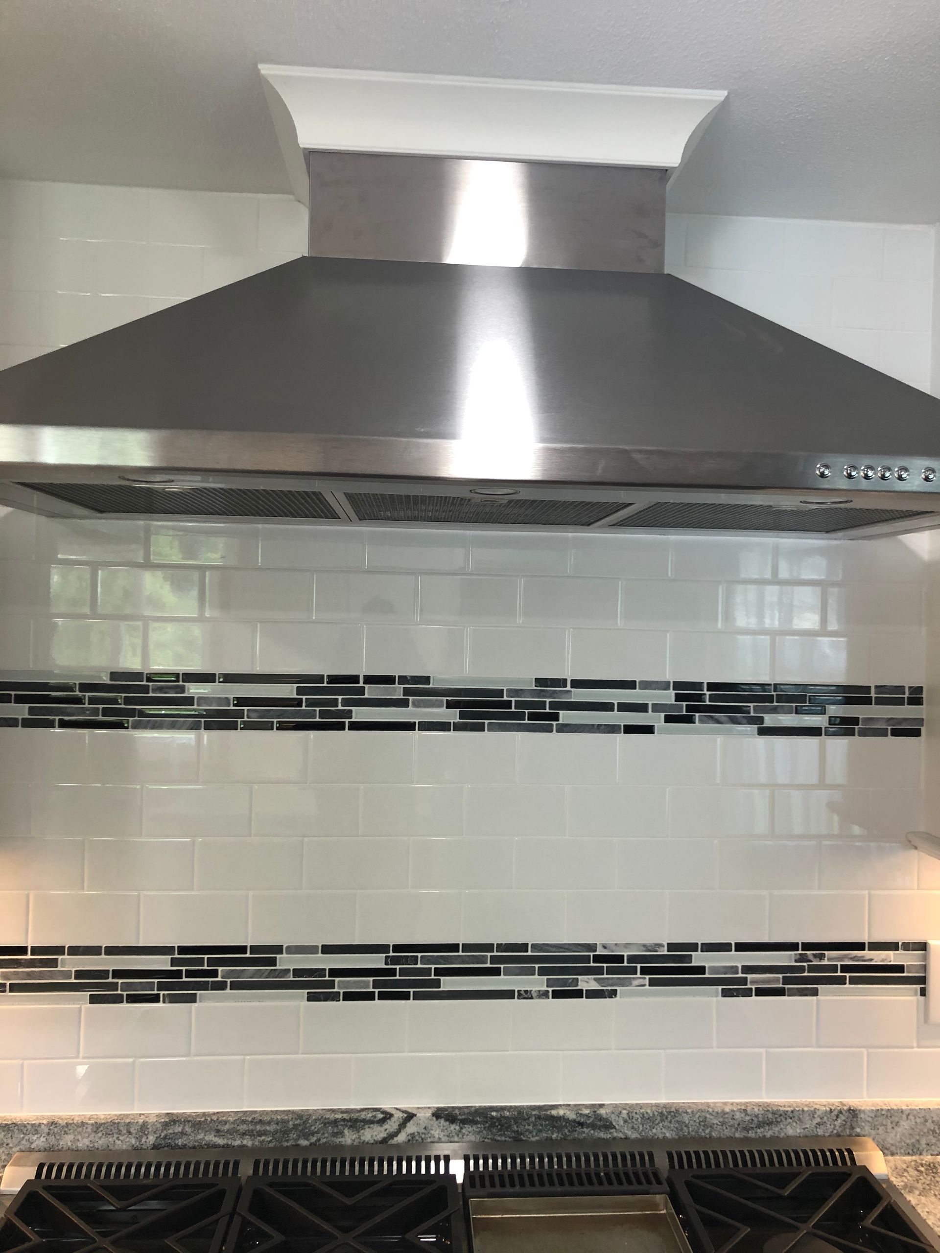 Stainless steel kitchen range hood above a white tile backsplash with black accent strips
