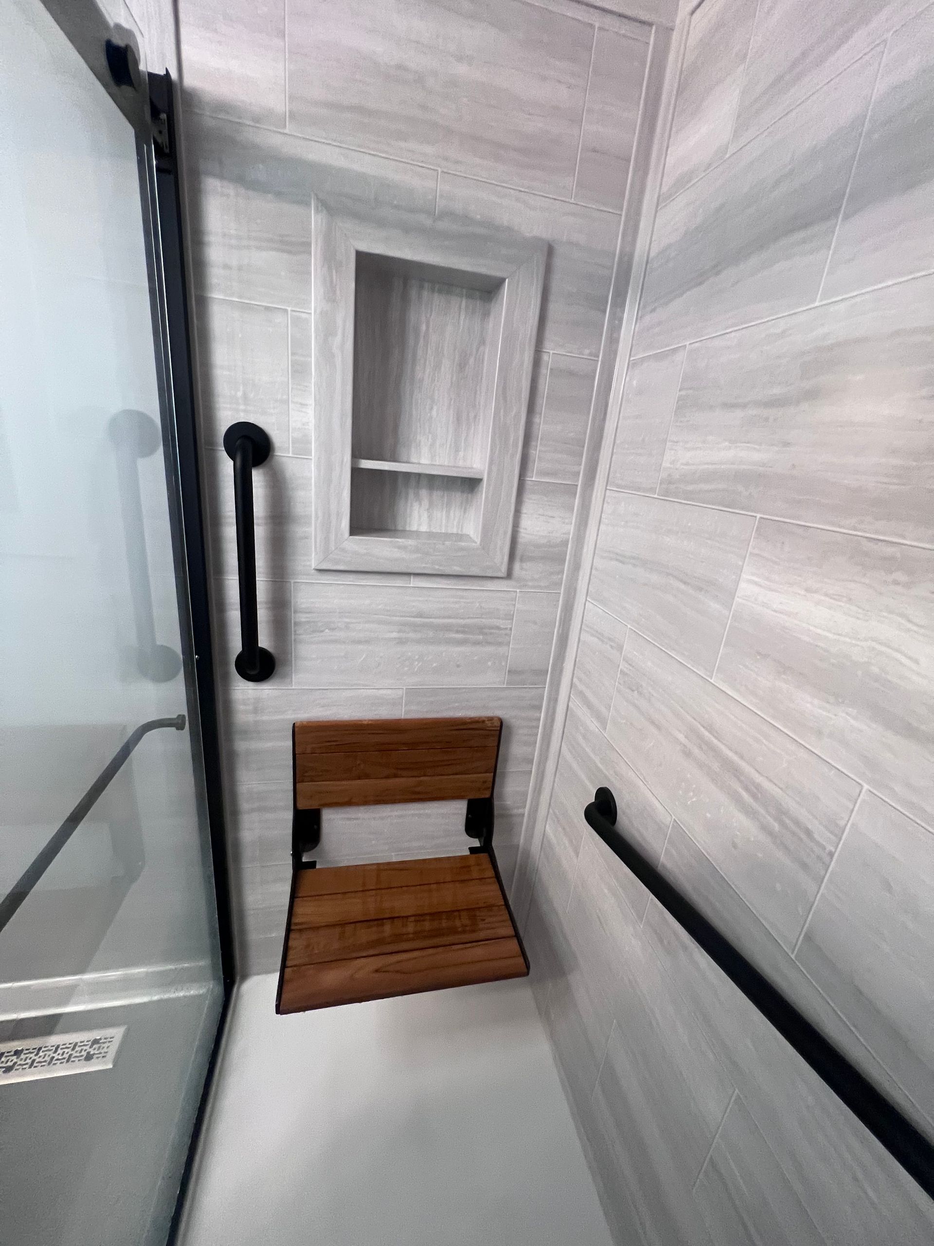 Narrow spa shower room with glass door, black grab bars, and a wooden seat against tiled walls