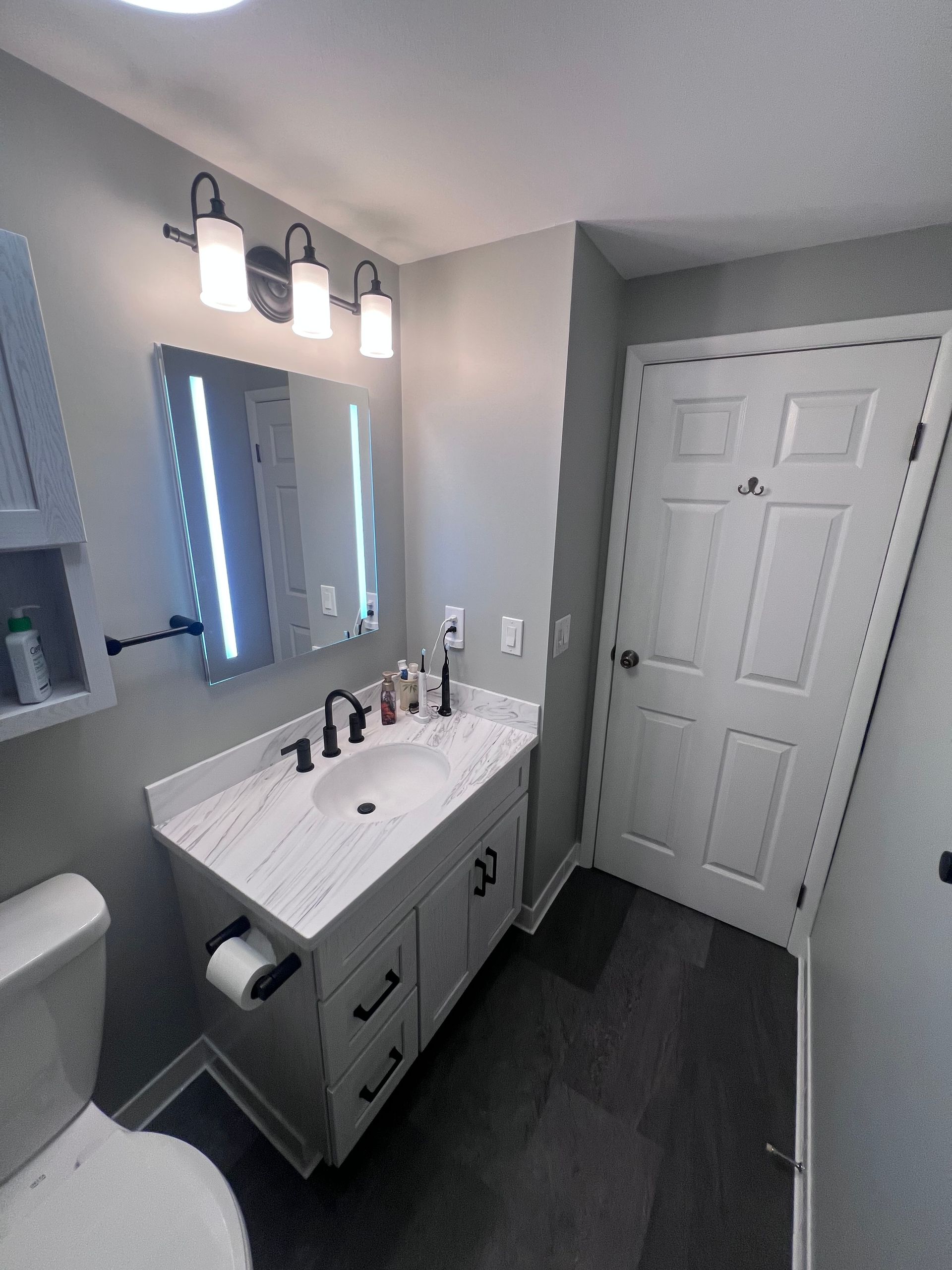 Small bathroom with vanity, mirror lights, toilet, and white door, shown from the doorway.