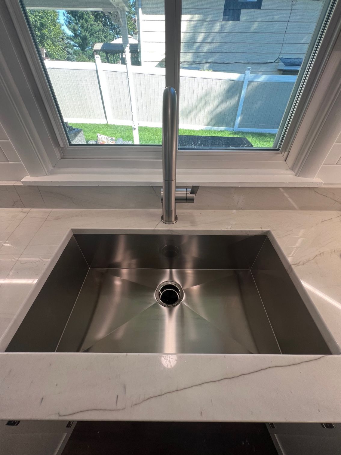 Stainless steel kitchen sink beneath a window, with a chrome faucet and white marble countertop.