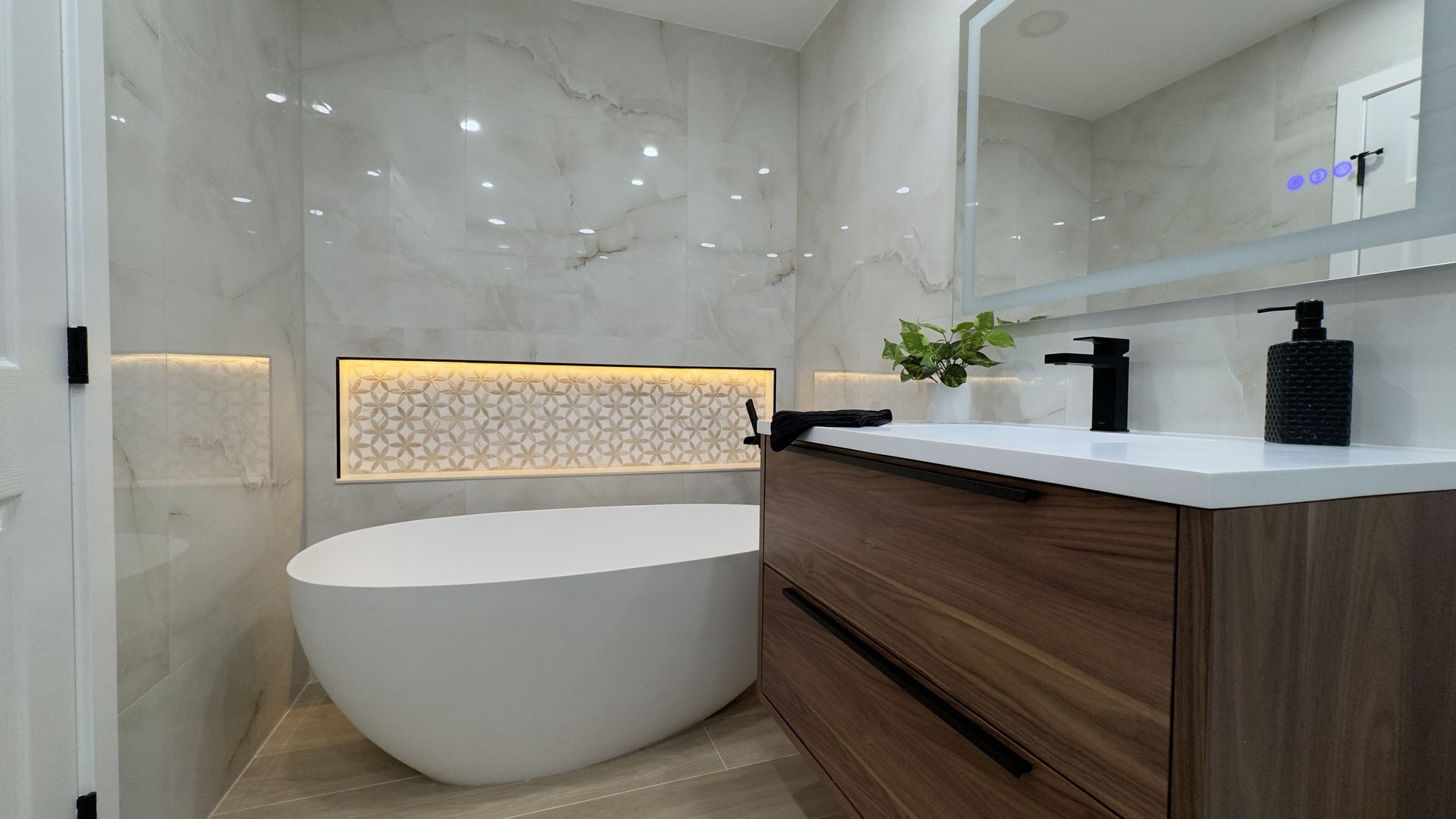 Modern bathroom with white tub, wood vanity, marble walls, and black fixtures.