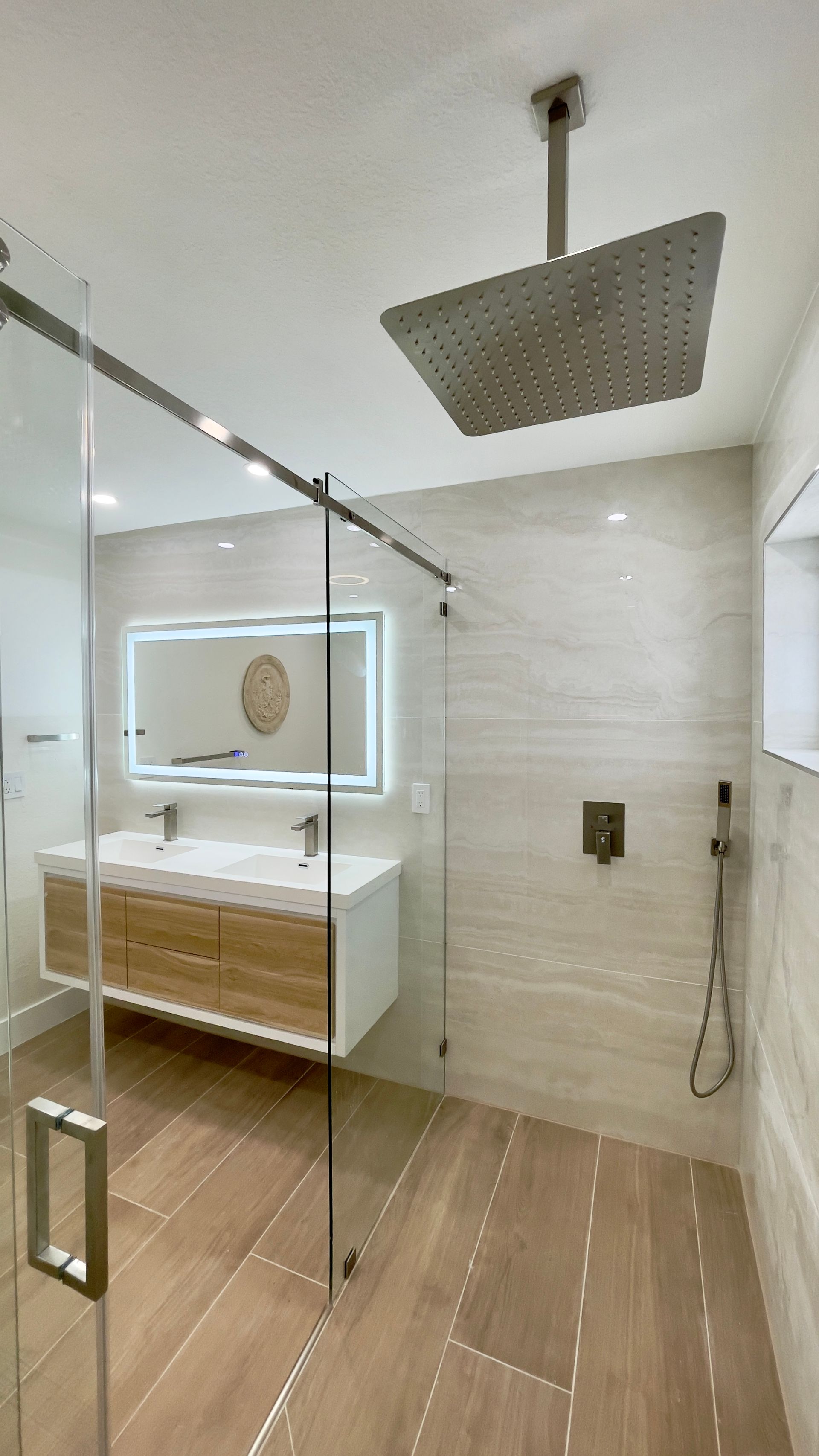 Modern bathroom with glass shower door, rain shower head, double sink vanity, and wood-look tile.
