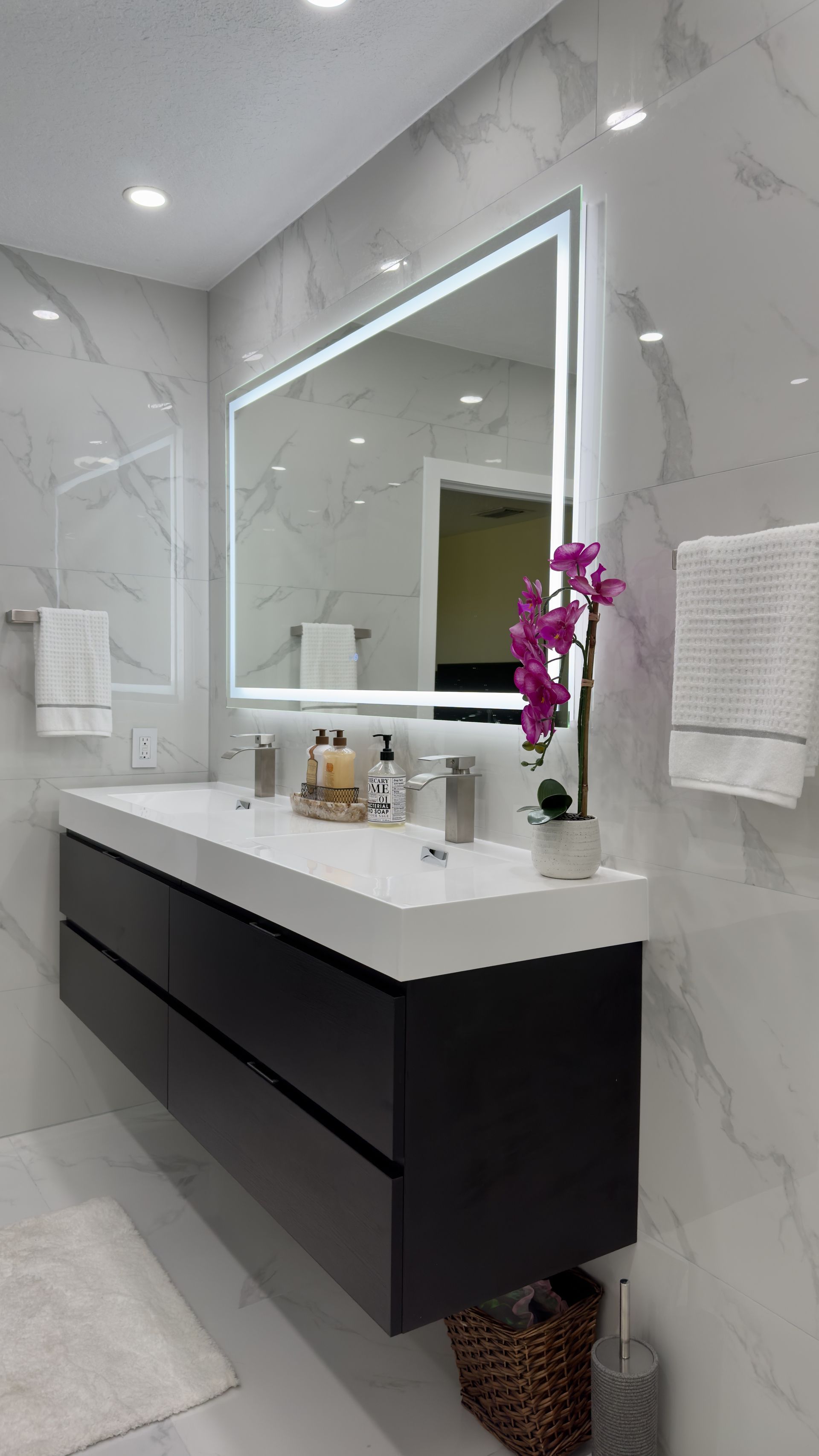 Modern bathroom with floating dark vanity, lit mirror, white marble walls.