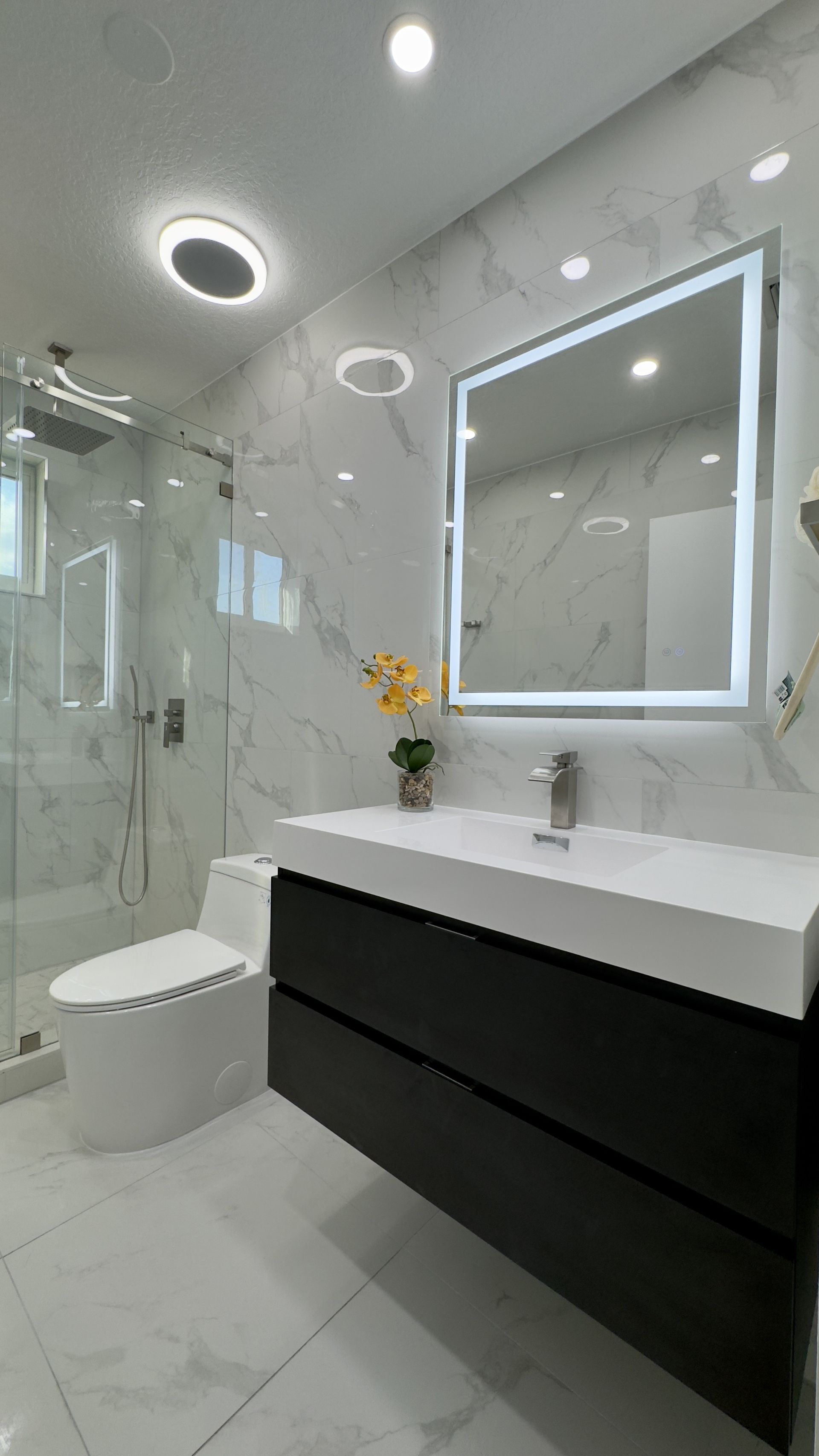 Modern bathroom with marble-like walls, a black vanity, and a lit-up mirror.