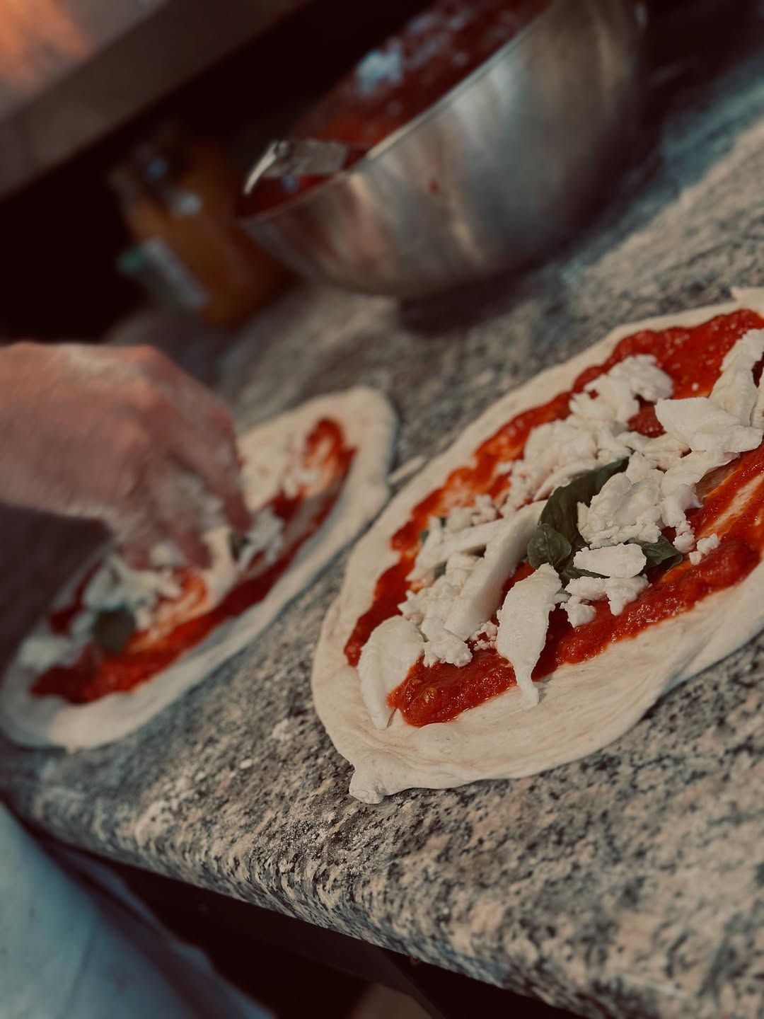 Due pizze preparate con salsa rossa, mozzarella e basilico su un bancone di marmo; i condimenti vengono aggiunti a mano.