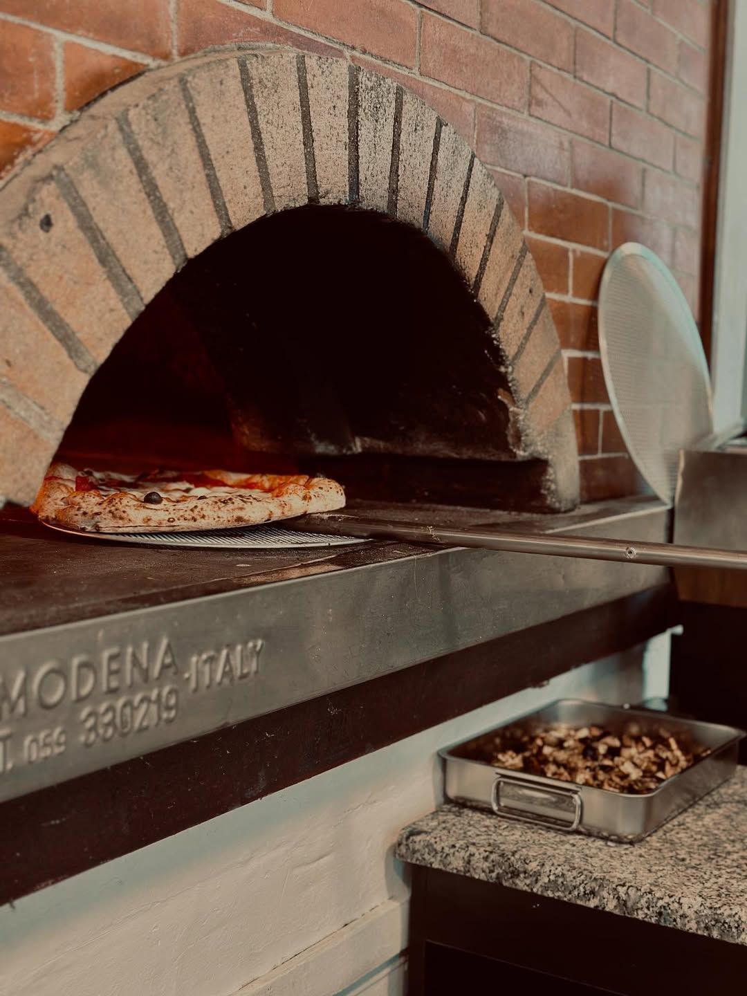 Pizza estratta da un forno a legna con una pala di metallo; forno di Modena, Italia.