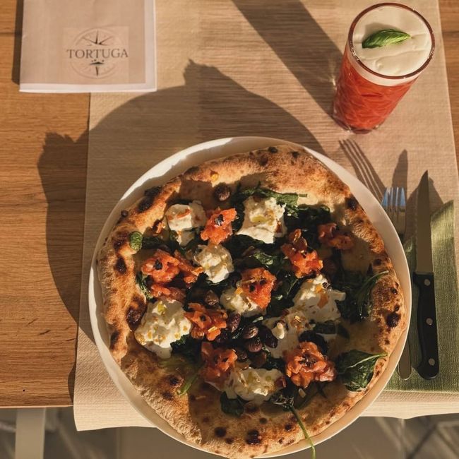 Pizza condita con verdure e formaggio, accanto a una bevanda rossa e posate su un tavolo del ristorante Tortuga.