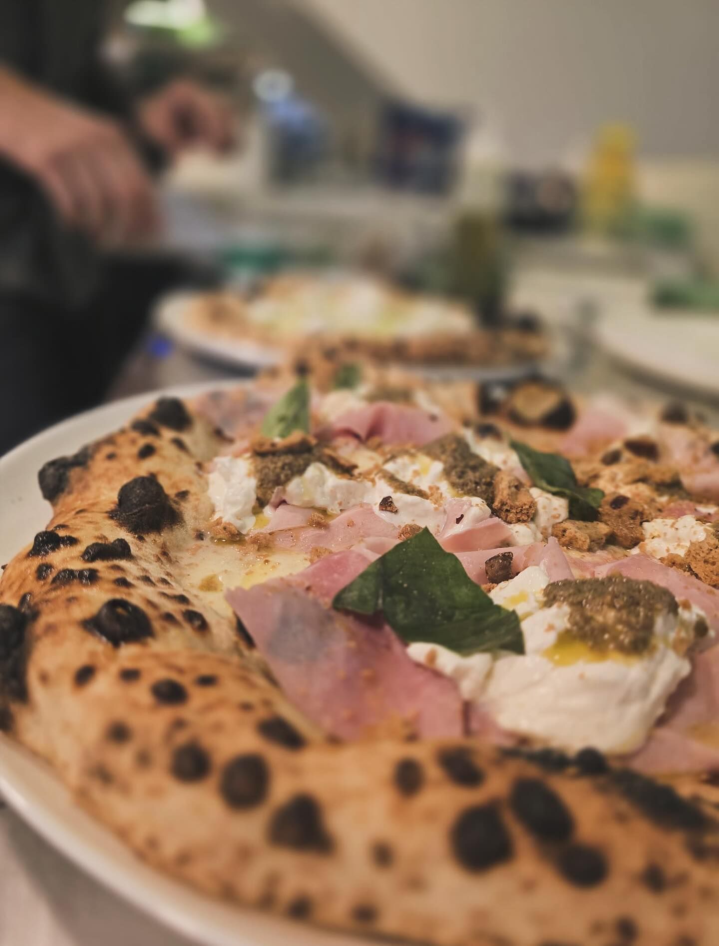 Primo piano di una pizza con crosta gonfie, farcita con prosciutto, formaggio, pesto e basilico.