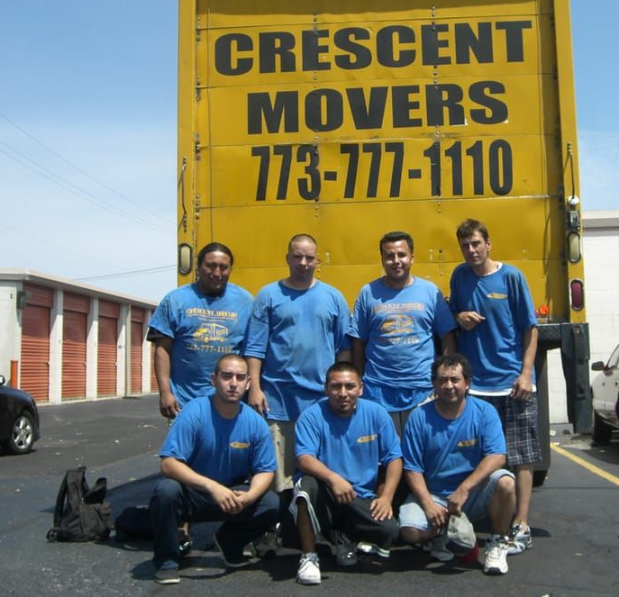 Movers in blue shirts pose in front of a yellow moving truck; storage units in background.