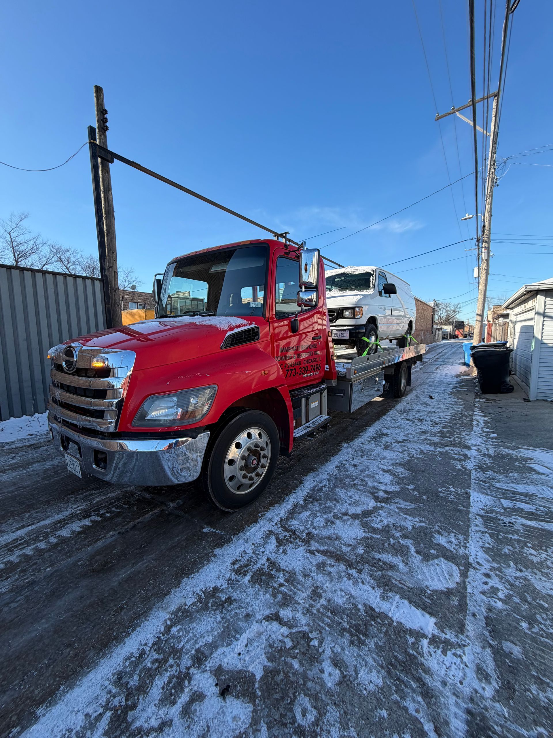 Red tow truck with a white van on its flatbed, parked on a snow-covered road. Bright sky in background.