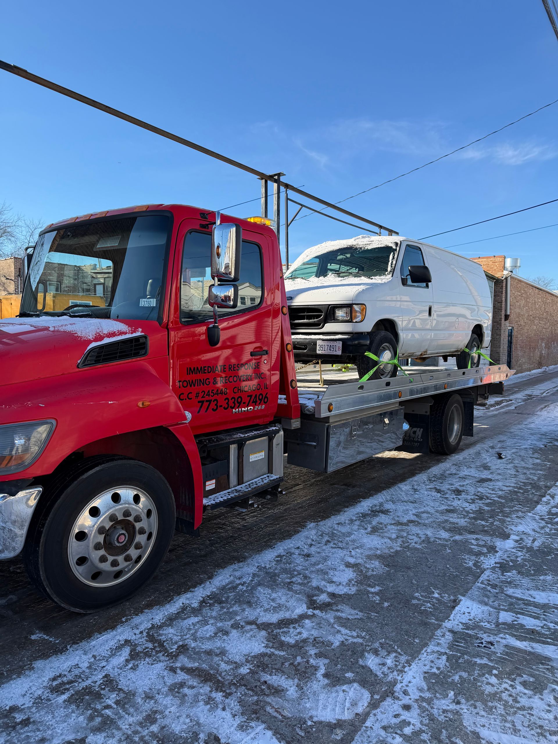 Red tow truck transporting a white van on a snowy street under a bright blue sky.