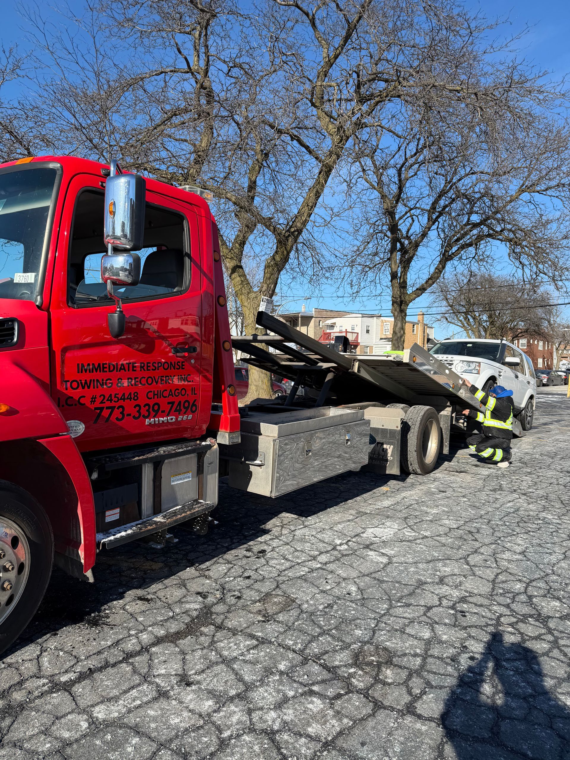 Red tow truck loading a white car on a sunny day. A person in a yellow vest assists.