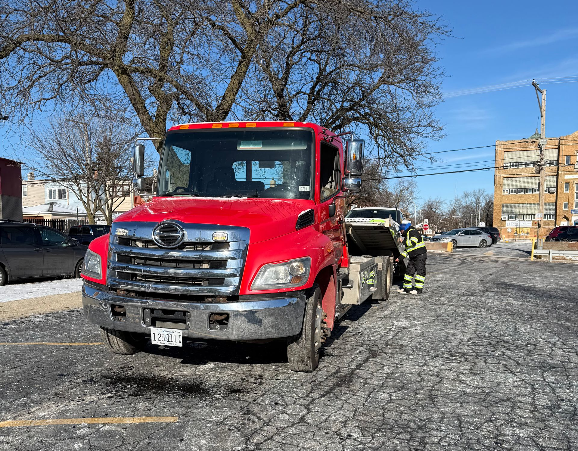 Red tow truck with a car on the bed, in a parking lot. Two people in firefighter gear. Bright, sunny day.