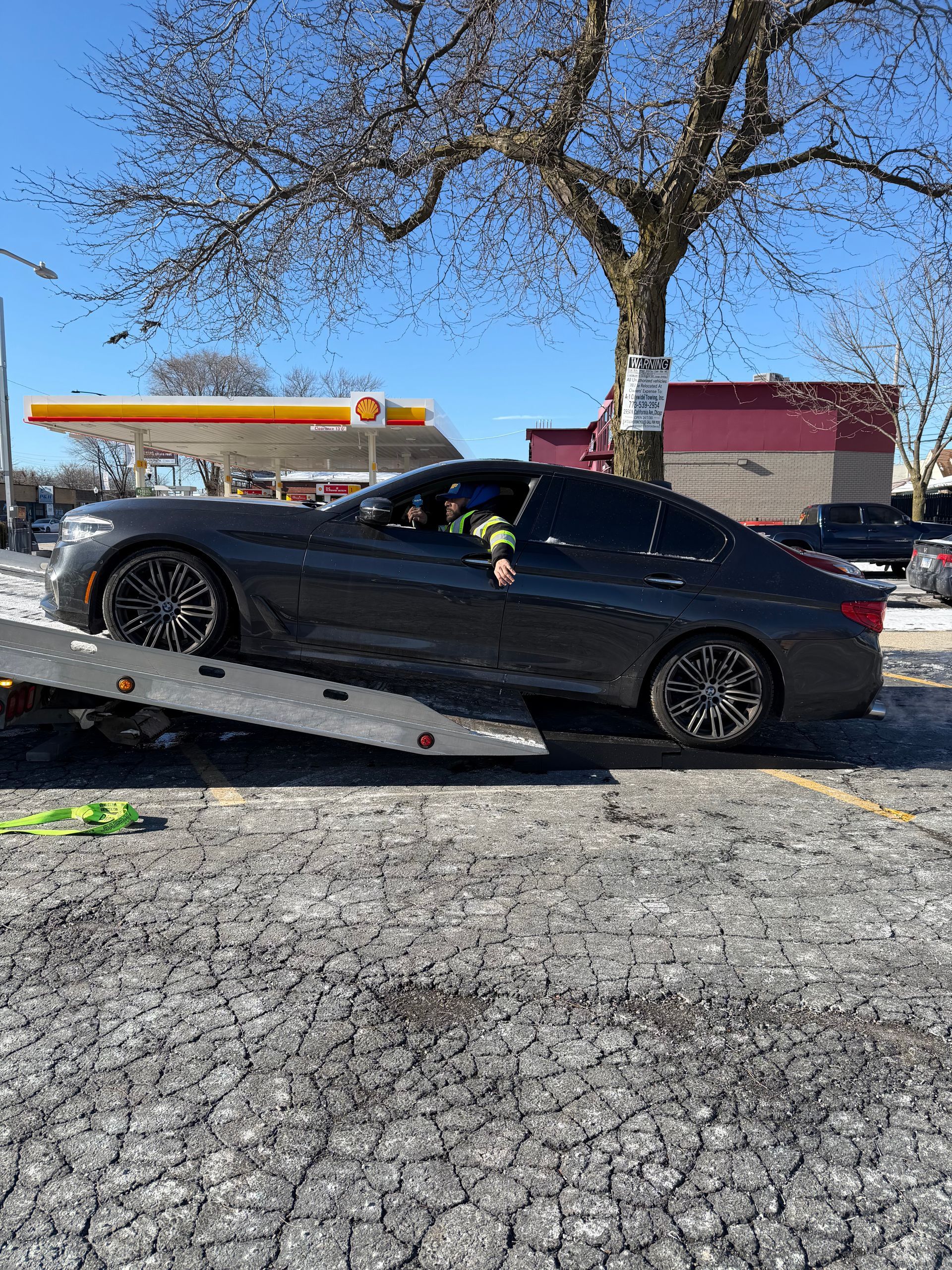 Gray car on a tow truck, near a Shell gas station. Clear sky, sunny day.