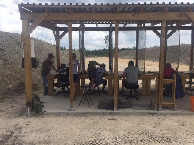 People at a shooting range, under a wooden shelter, shooting guns. Cloudy day.
