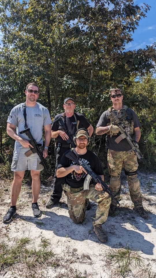 Four armed men pose outdoors. One kneels, holding a rifle. Others stand, holding rifles, wearing casual attire.