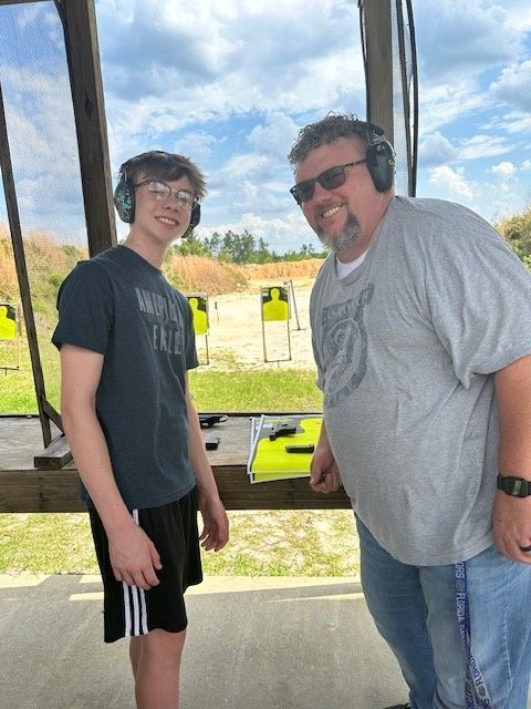 Man and teen at a shooting range, both wearing ear protection. Smiling, sunny day, targets in the background.