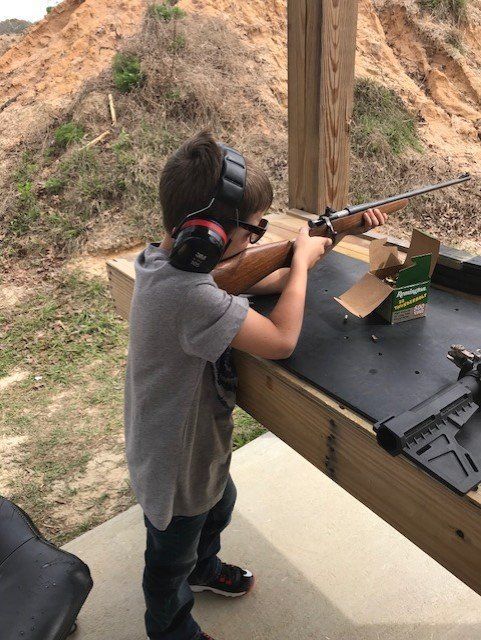 Boy aiming a rifle at a shooting range, wearing hearing protection and glasses.