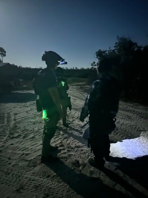 Three figures in tactical gear stand on a sandy path at night, illuminated by green and white lights.