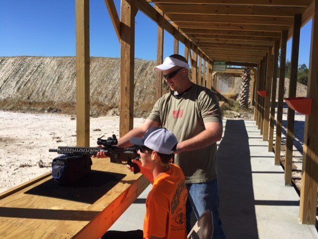 Man and child at a shooting range. Man shows child how to aim a rifle, outdoors on a sunny day.