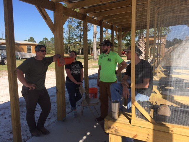 Four people stand under a wooden shelter. One leans, three converse. Sunny, outdoor setting.