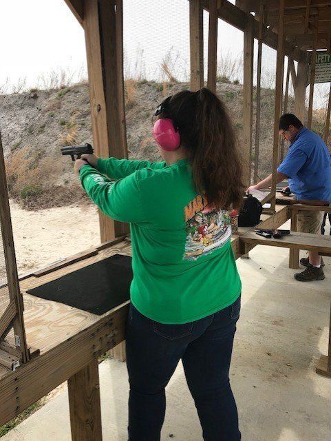 Woman in green shirt and pink earmuffs fires a handgun at an outdoor shooting range.