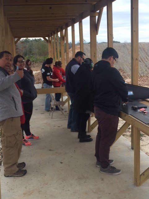 People shooting guns at an outdoor shooting range. Some are standing, others aiming. Cloudy day.