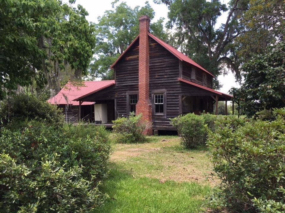 Two-story wooden house with a red roof and chimney, surrounded by green bushes and trees.