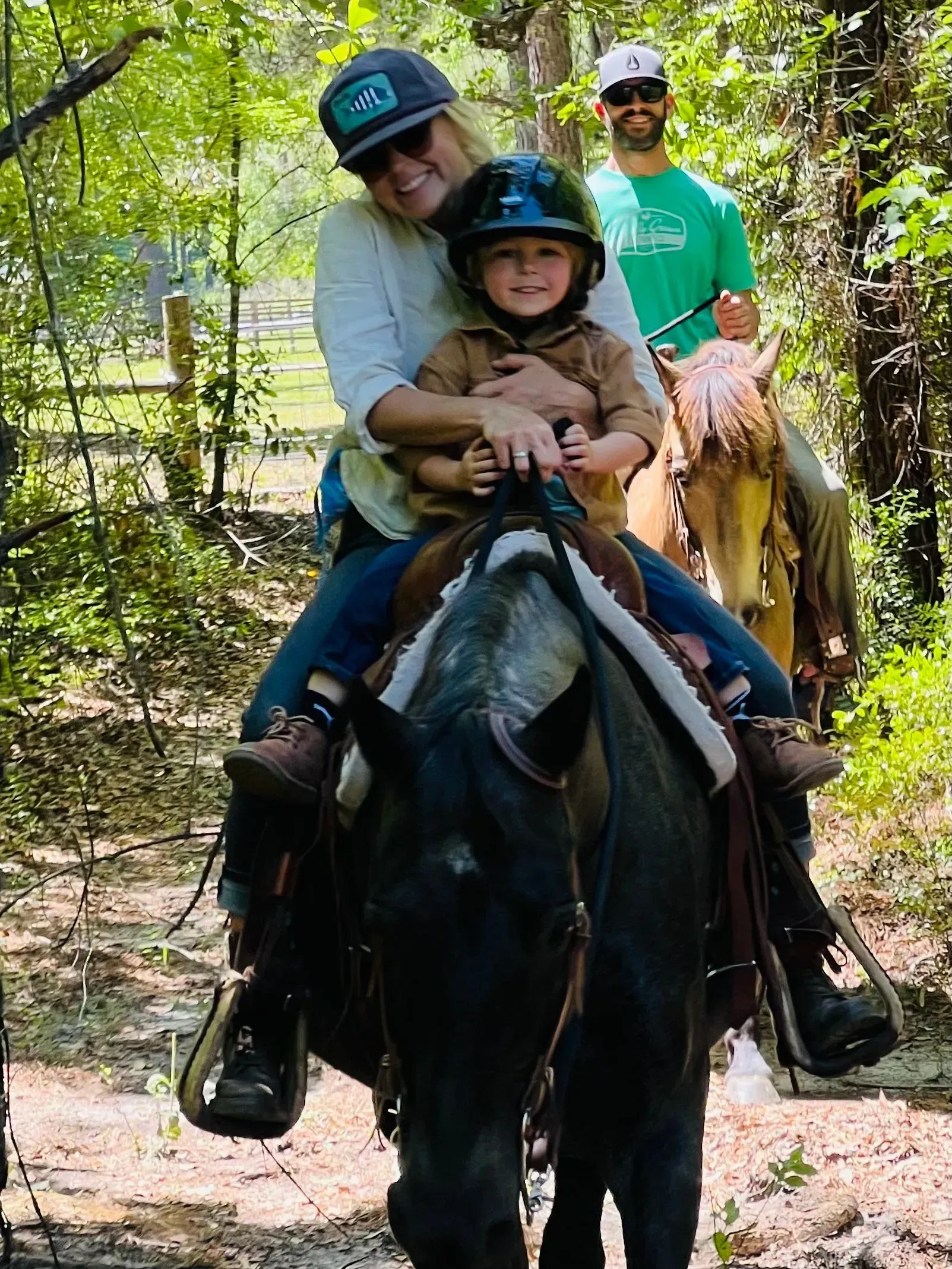 Woman and child horseback riding; man rides behind in woods.