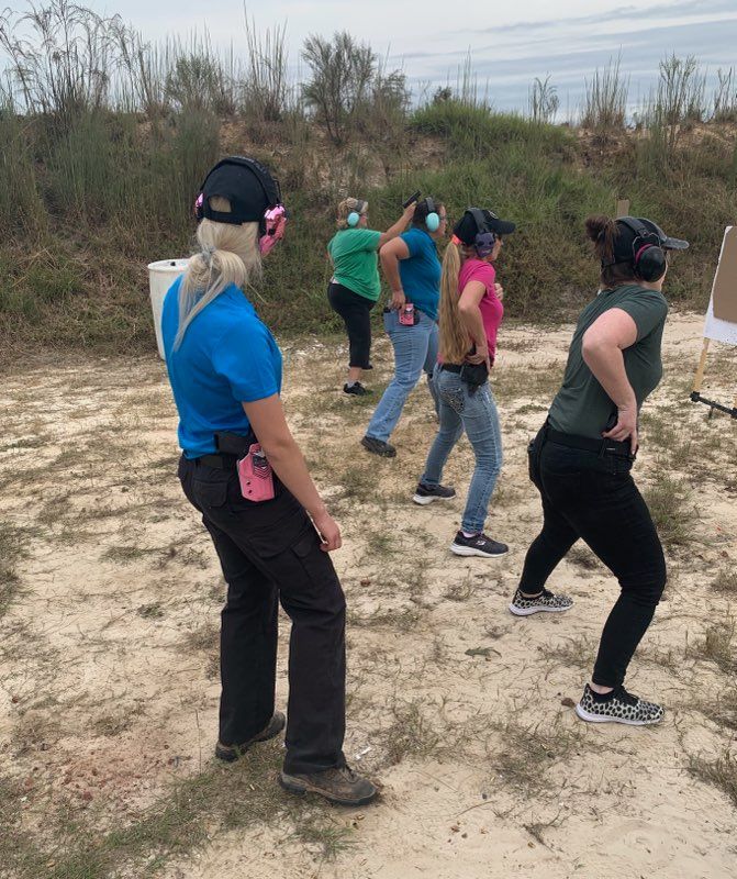 Women practice drawing pistols at an outdoor shooting range. They wear ear protection, aiming in the same direction.