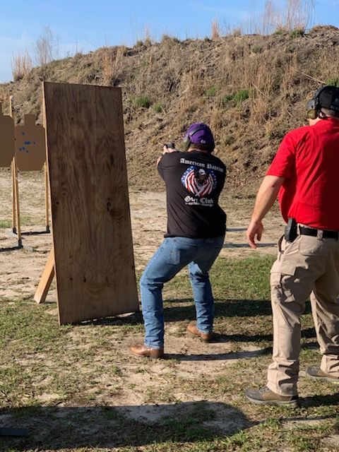 Person in purple hat shoots a gun at a target. Man in red shirt watches. Outdoor shooting range.