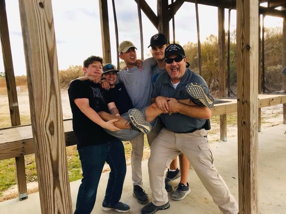 Men smiling and posing playfully at an outdoor shooting range, holding each other up.