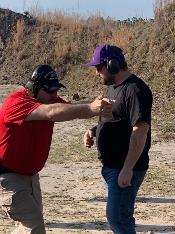 Two men at a shooting range, one in red shirt aiming a gun, other in black shirt and purple hat watching.