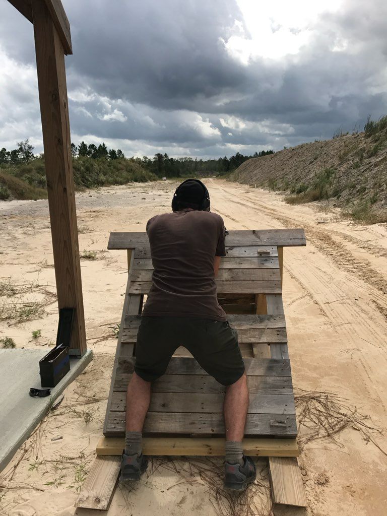 Man shooting a gun, aiming over a wooden structure at an outdoor shooting range. Cloudy sky overhead.