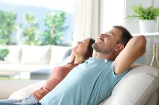 Two people relaxing on a sofa in a bright living room, leaning back with eyes closed.