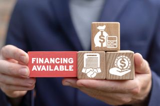 Hands holding wooden blocks and a red block reading “FINANCING AVAILABLE” against a blurred suit background