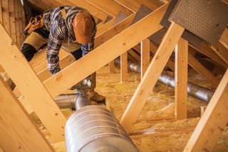 Worker crouching in an attic, inspecting ductwork and wooden beams above insulation