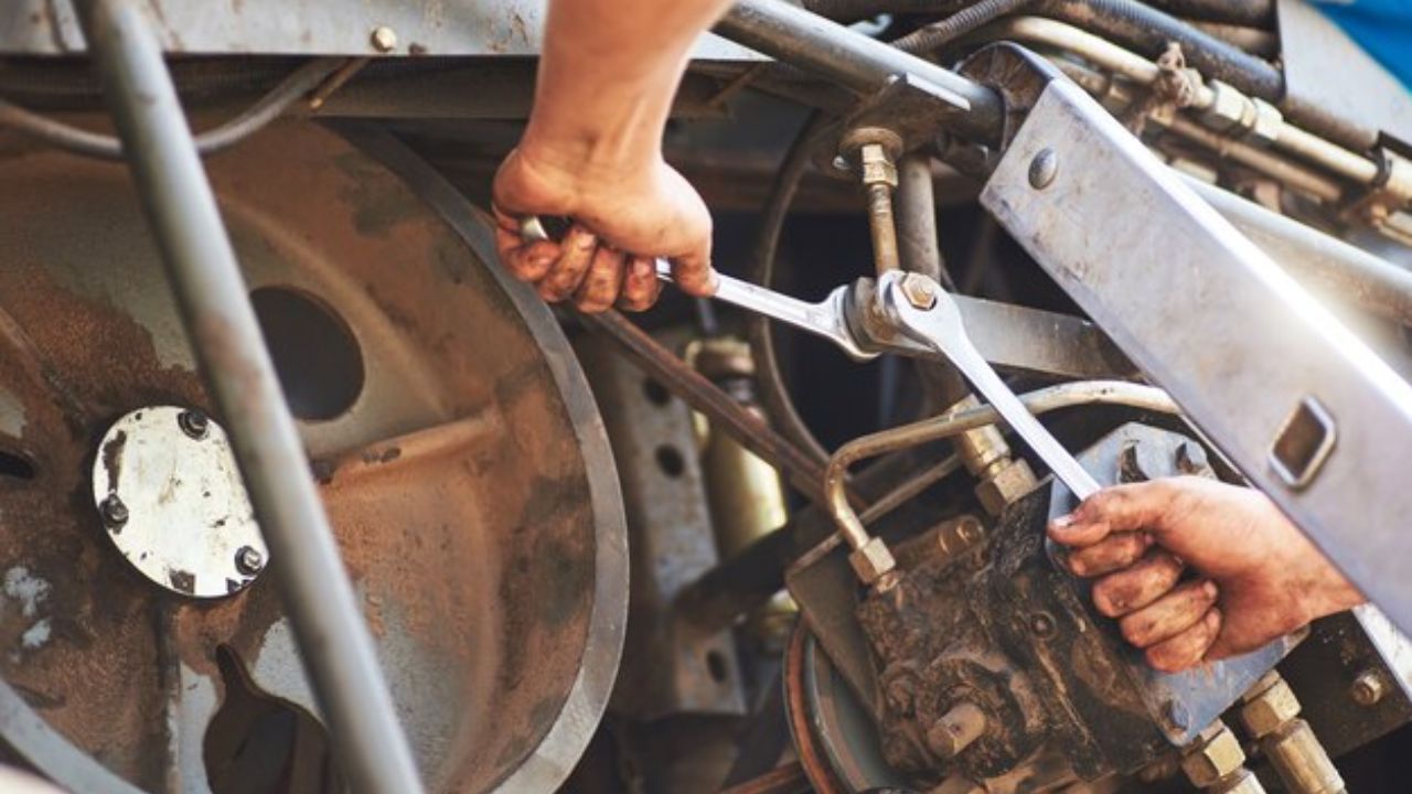  Mechanic tightening bolts with wrenches while repairing heavy machinery with belts, pulleys, and hydraulic components visible.