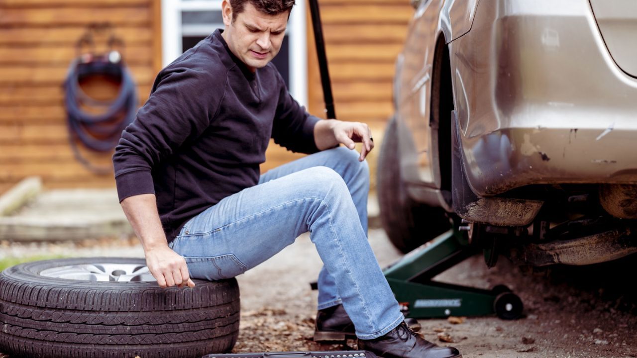 Man kneeling beside a car lifted on a jack, checking a tire with a socket wrench 
