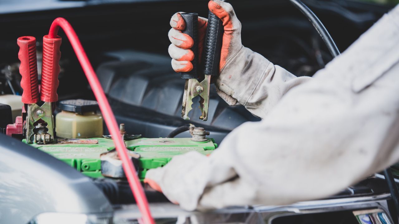 Close-up of gloved hands attaching jumper cable clamps to a car battery terminal under the hood
