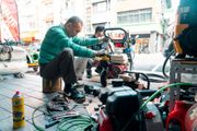 Mechanic kneels on a sidewalk repairing equipment beside tools and gas cans in a city street
