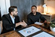 Two colleagues in a meeting, reviewing documents at a desk with a laptop and charts.