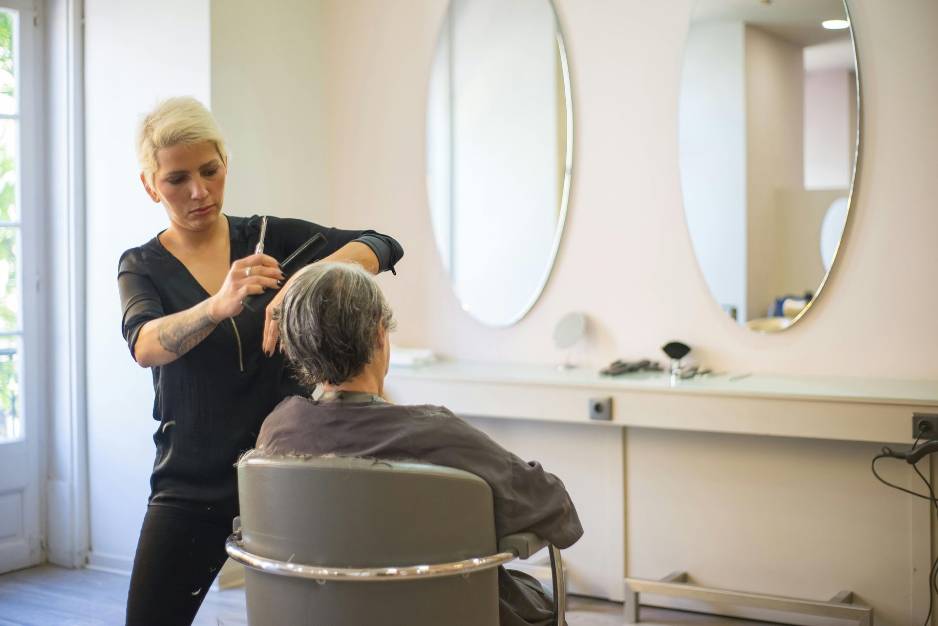 Hairdresser cutting a client’s hair in a bright salon with mirrors and a window.