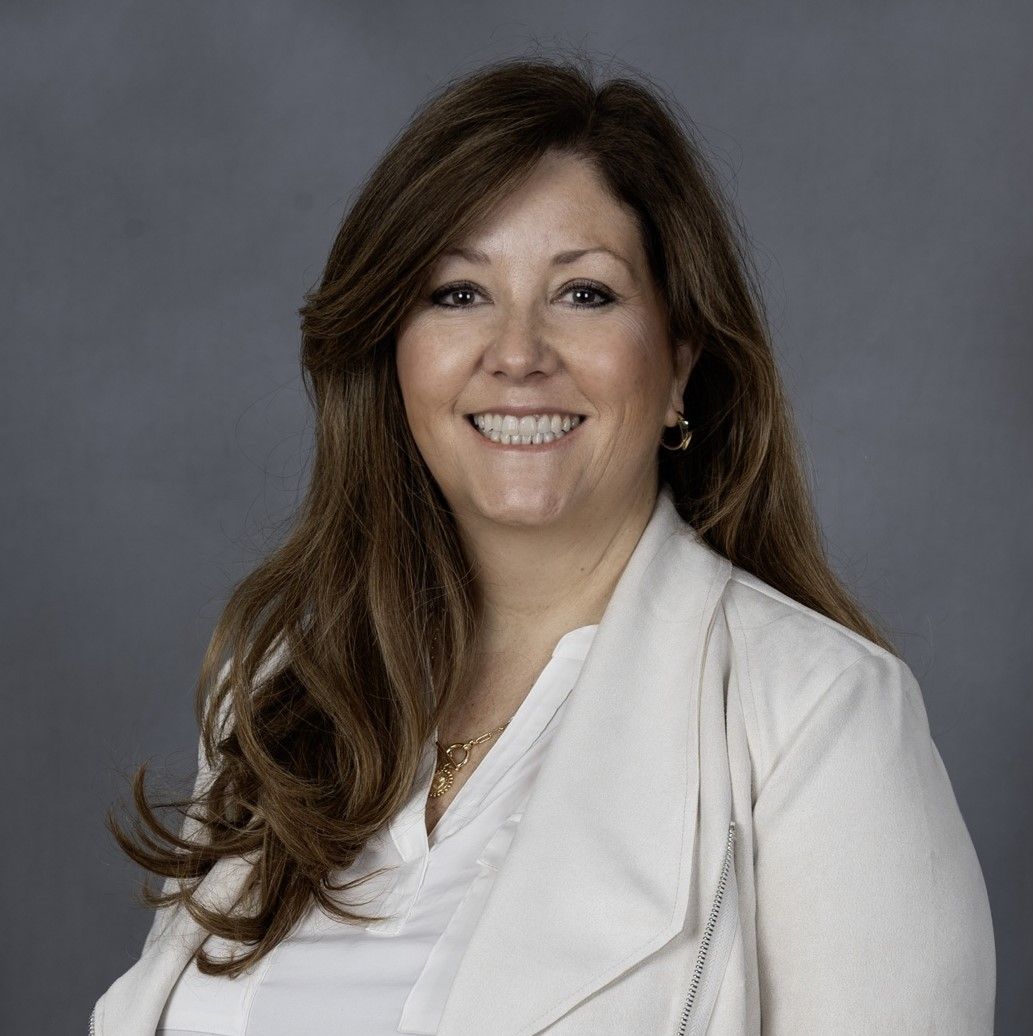 Woman with long brown hair smiling, wearing a white jacket and shirt, against a gray background.