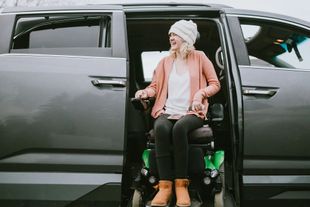 Woman in wheelchair exiting accessible van, smiling. She wears a pink jacket, white beanie and boots.