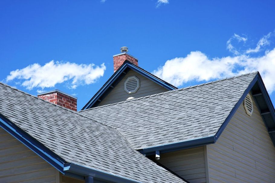 A residential house with gray shingled roofs, two brick chimneys, and tan siding against a bright blue sky with clouds.