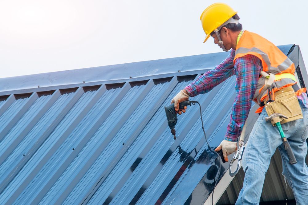 A worker wearing a yellow hard hat and orange safety vest drills a fastener into a corrugated metal roof.