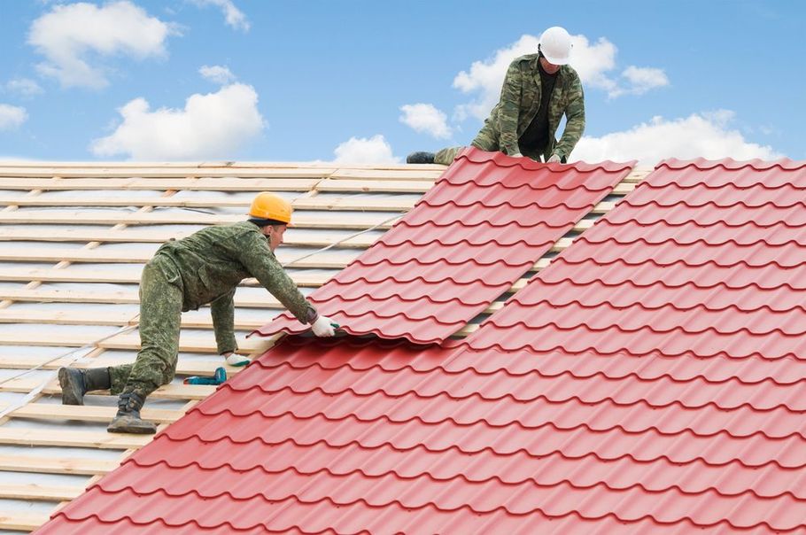 Two people in camouflage workwear install red corrugated metal roofing sheets on a wooden roof frame against a blue sky.